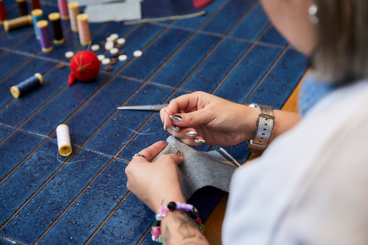 Close-up of a person hand-stitching a piece of denim on a blue worktable, with spools of thread, scissors, buttons and a red pin cushion scattered nearby 