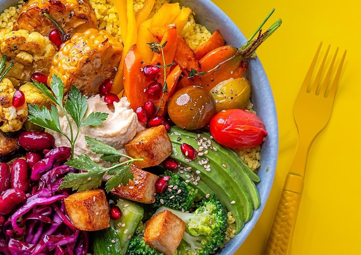 Colorful vegan grain bowl with roasted vegetables, tofu, hummus, avocado and pomegranate seeds on a yellow background 
