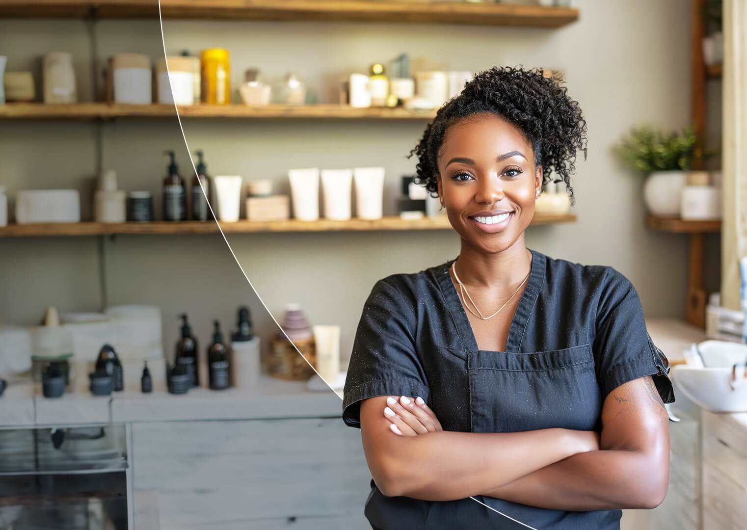 A beauty professional stands smiling with arms crossed in a well-organized studio, surrounded by neatly arranged skincare and haircare products on wooden shelves 