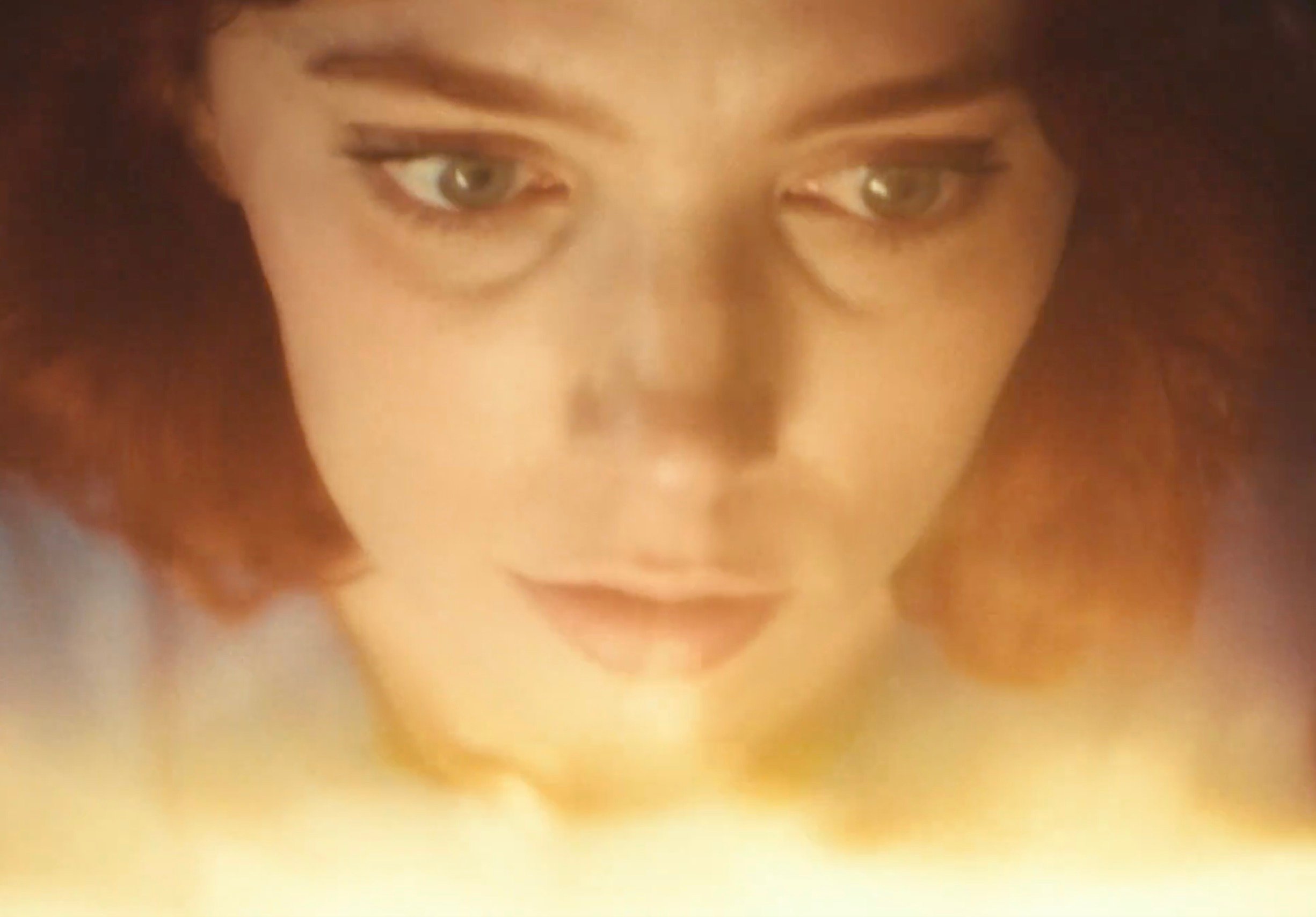 A close-up of a person with red hair gazing downward, their face softly lit by a warm golden glow that reflects in their eyes, creating an intense, absorbed expression 