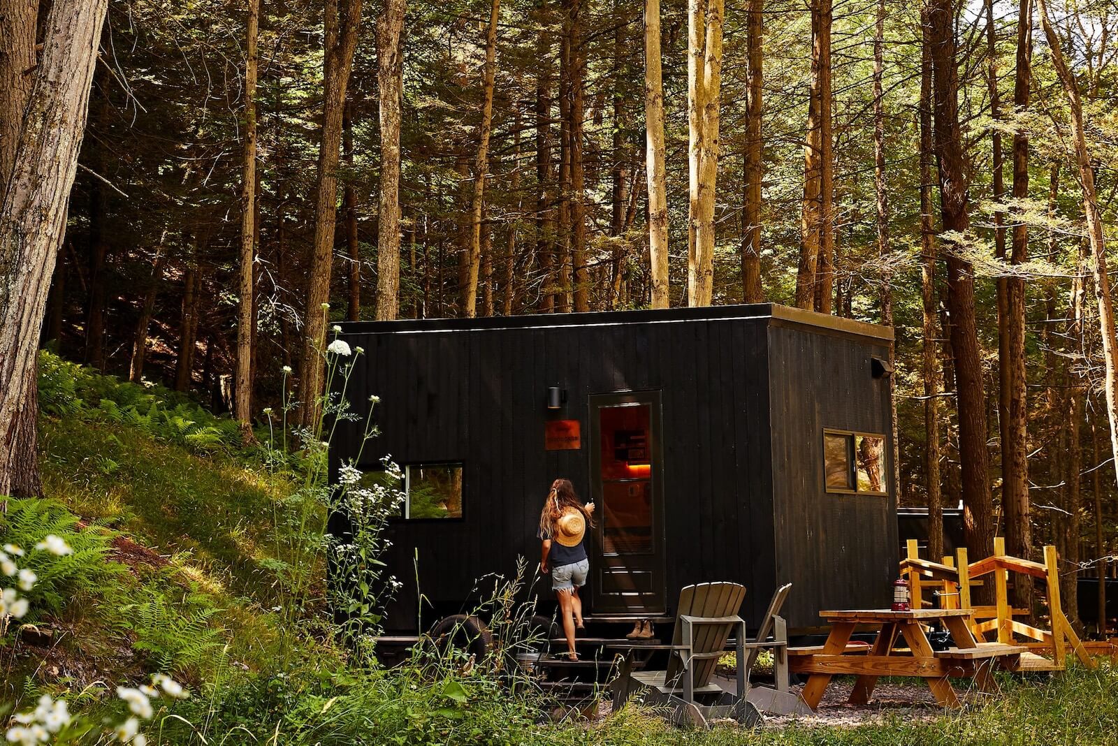A woman with a straw hat approaches a small black cabin surrounded by tall trees, with outdoor seating and a picnic table in the forest 