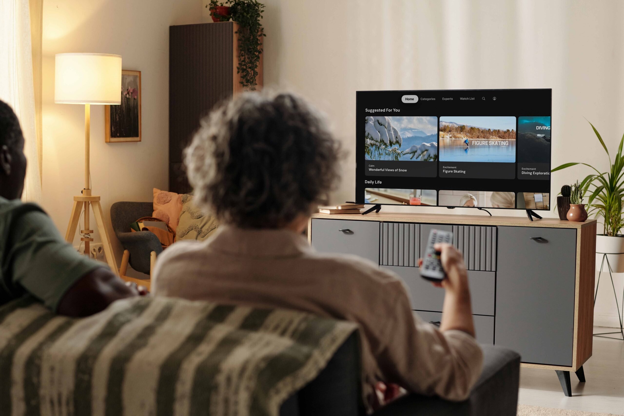 An older woman sits on a couch holding a remote control, watching a TV screen displaying the Menta streaming platform interface with categories including snowy nature scenes, figure skating and diving content 