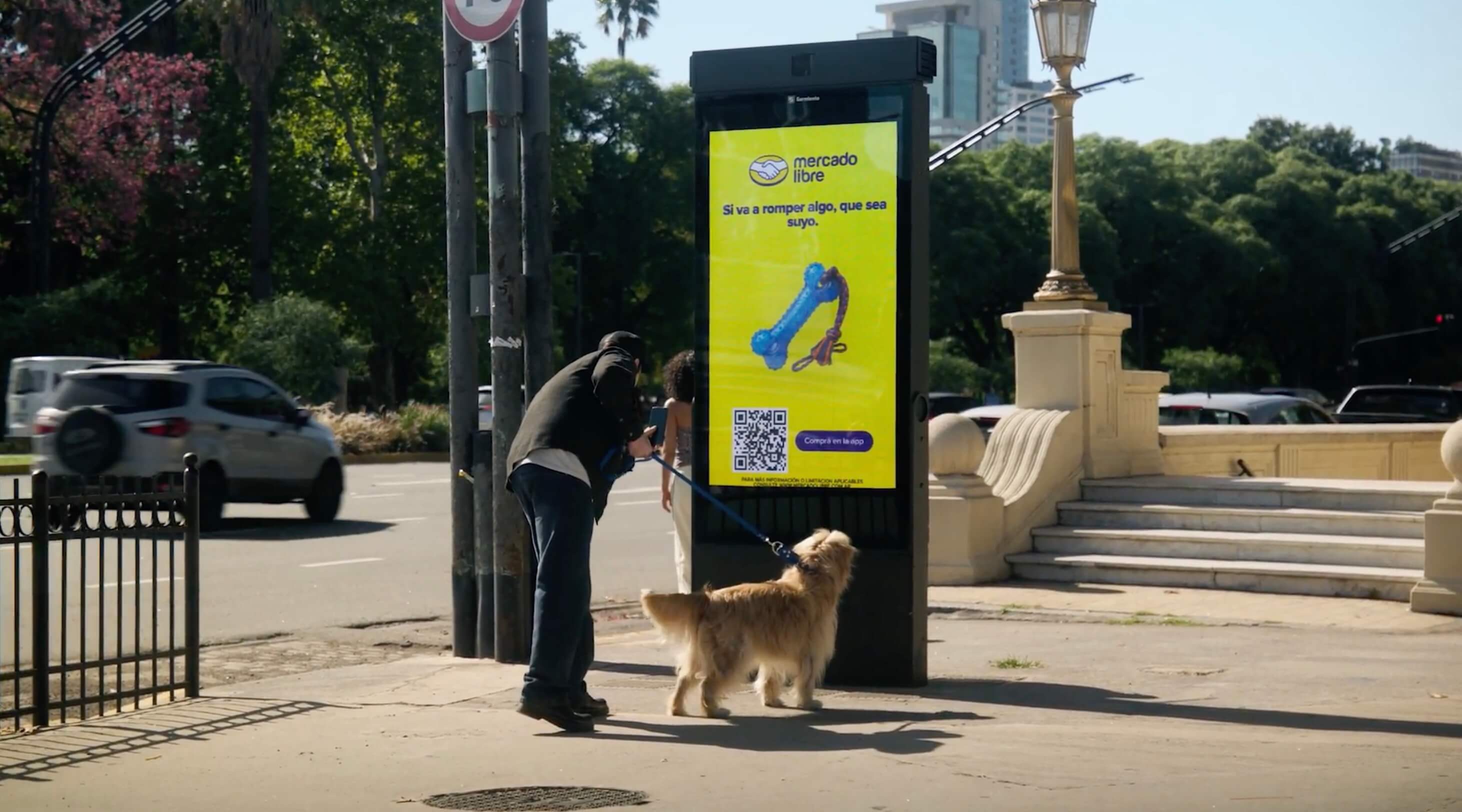 A person walking a dog on a leash pauses beside a Mercado Libre digital billboard on a Buenos Aires street. The yellow billboard displays a dog chew toy and rope with the Spanish text 