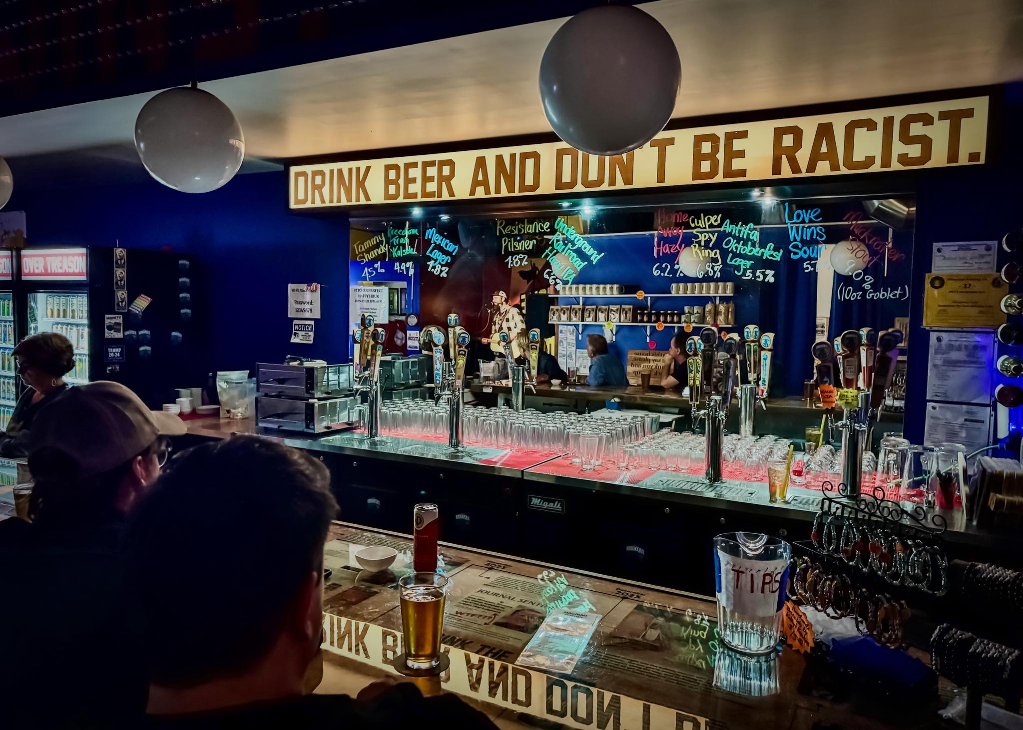 The bar at Minocqua Brewing Company's taproom, with beer taps, glasses and a handwritten chalkboard menu listing brews like Resistance Pilsner and Love Wins Sour, beneath a large sign reading 
