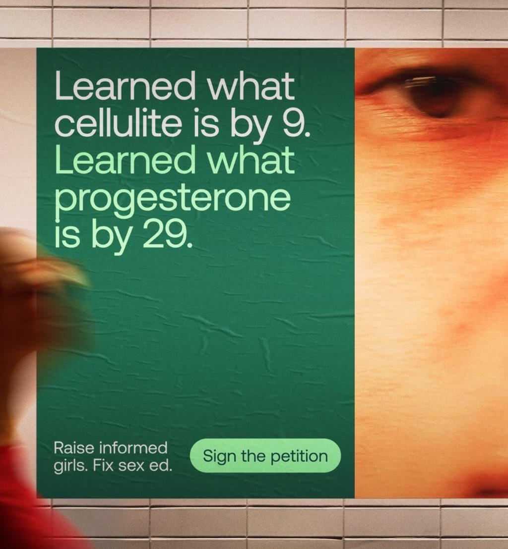 Poster on a tiled wall with green background text reading “Learned what cellulite is by 9. Learned what progesterone is by 29.” A blurred figure passes in front, and smaller text below reads “Raise informed girls. Fix sex ed. Sign the petition,” alongside a close-up of part of a person’s face 