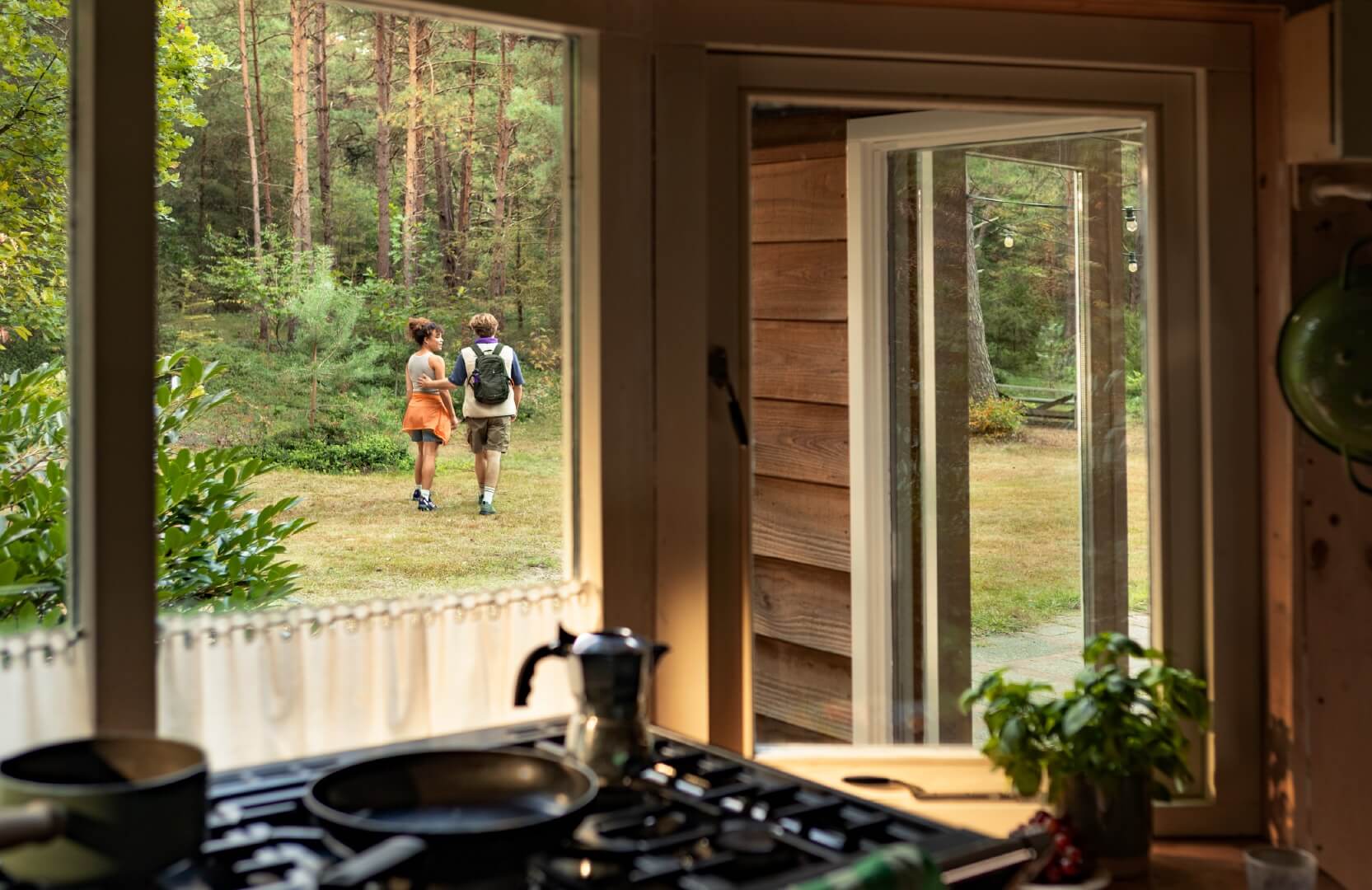 A view from inside a cozy cabin looking out through open doors to a couple walking together in a forest clearing, with the cabin's kitchen counter and stove in the foreground 