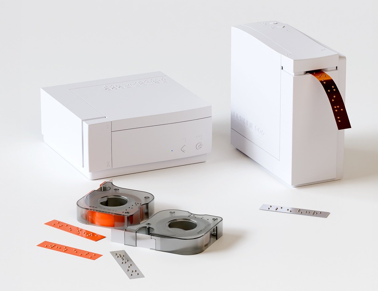 Two minimalist white label-printing devices on a light background, shown alongside transparent tape cartridges and narrow adhesive strips printed with raised Braille dots, one device actively dispensing a Braille label strip 