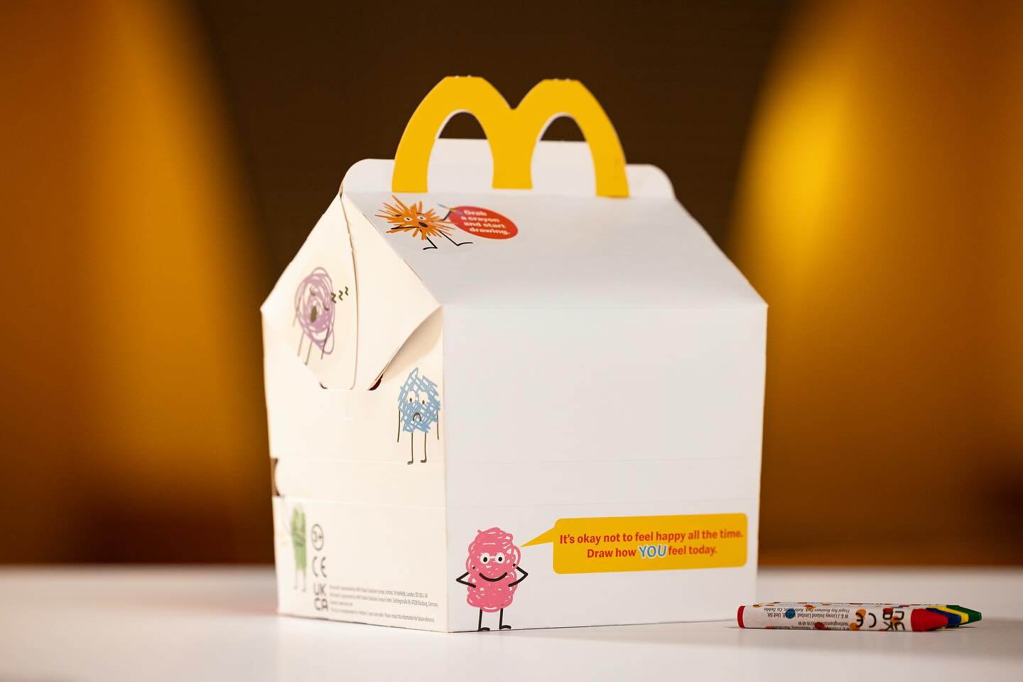 A McDonald’s Happy Meal box redesigned as a blank canvas, accompanied by the message “It’s okay not to feel happy all the time. Draw how YOU feel today.” A small pack of crayons lies next to the box on a white surface, against a warm yellow background 