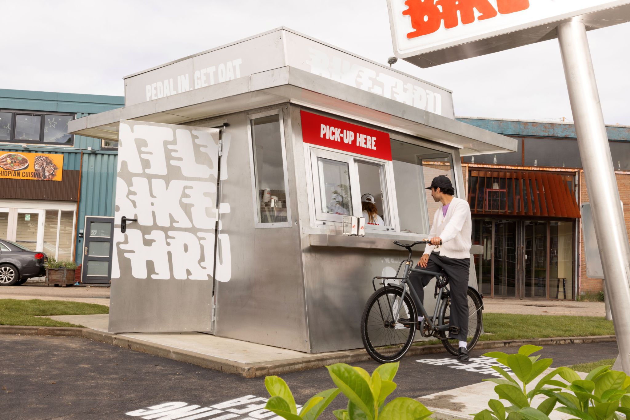 A cyclist on a black bike pulls up to the pick-up window of Oatly's stainless-steel Bike-Thru kiosk, marked with bold white 