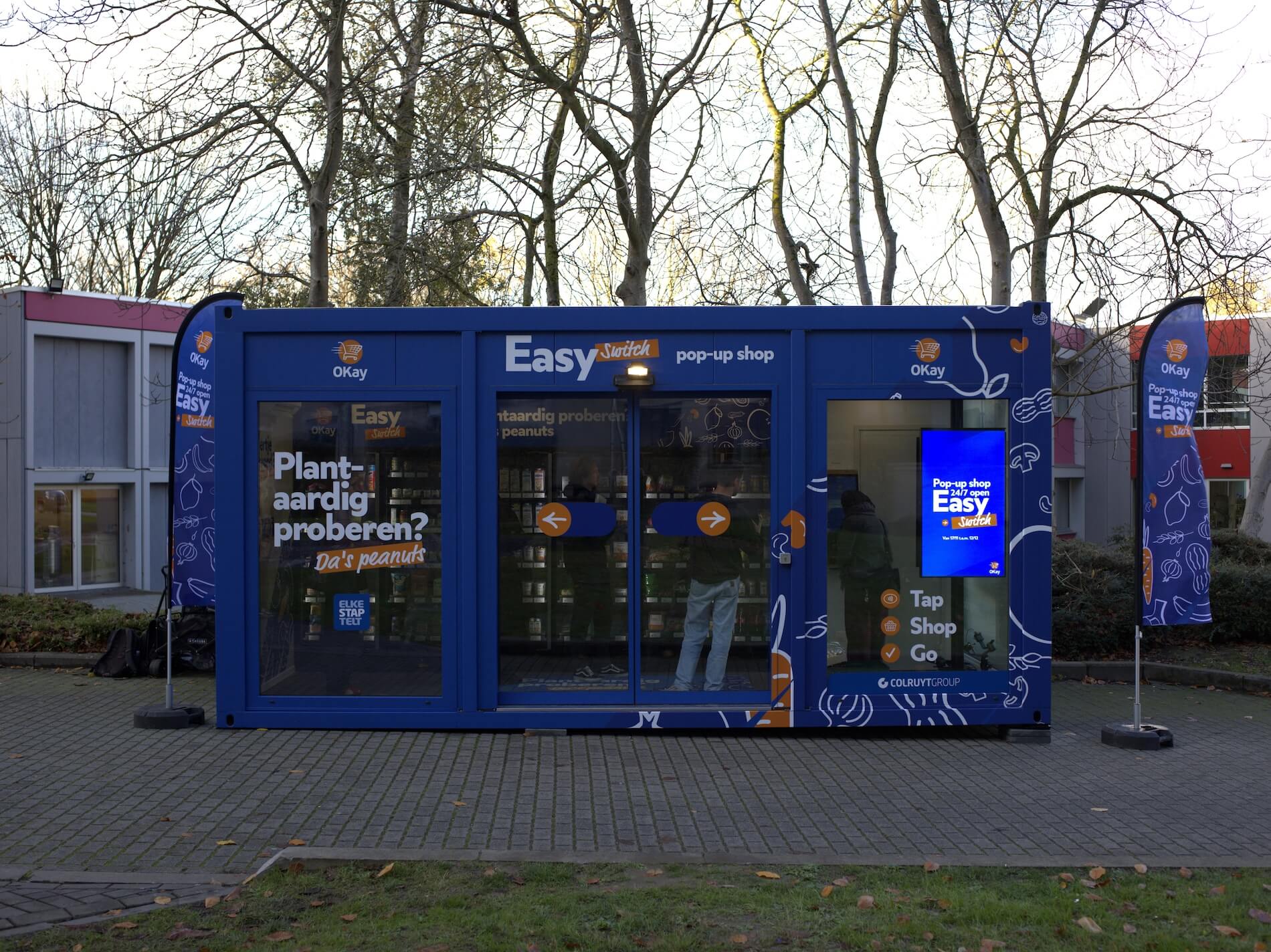 A blue modular pop-up grocery shop branded “Okay Easy Switch” sits outdoors on a paved walkway. The exterior displays text promoting plant-based products and 24/7 tap-and-go shopping 