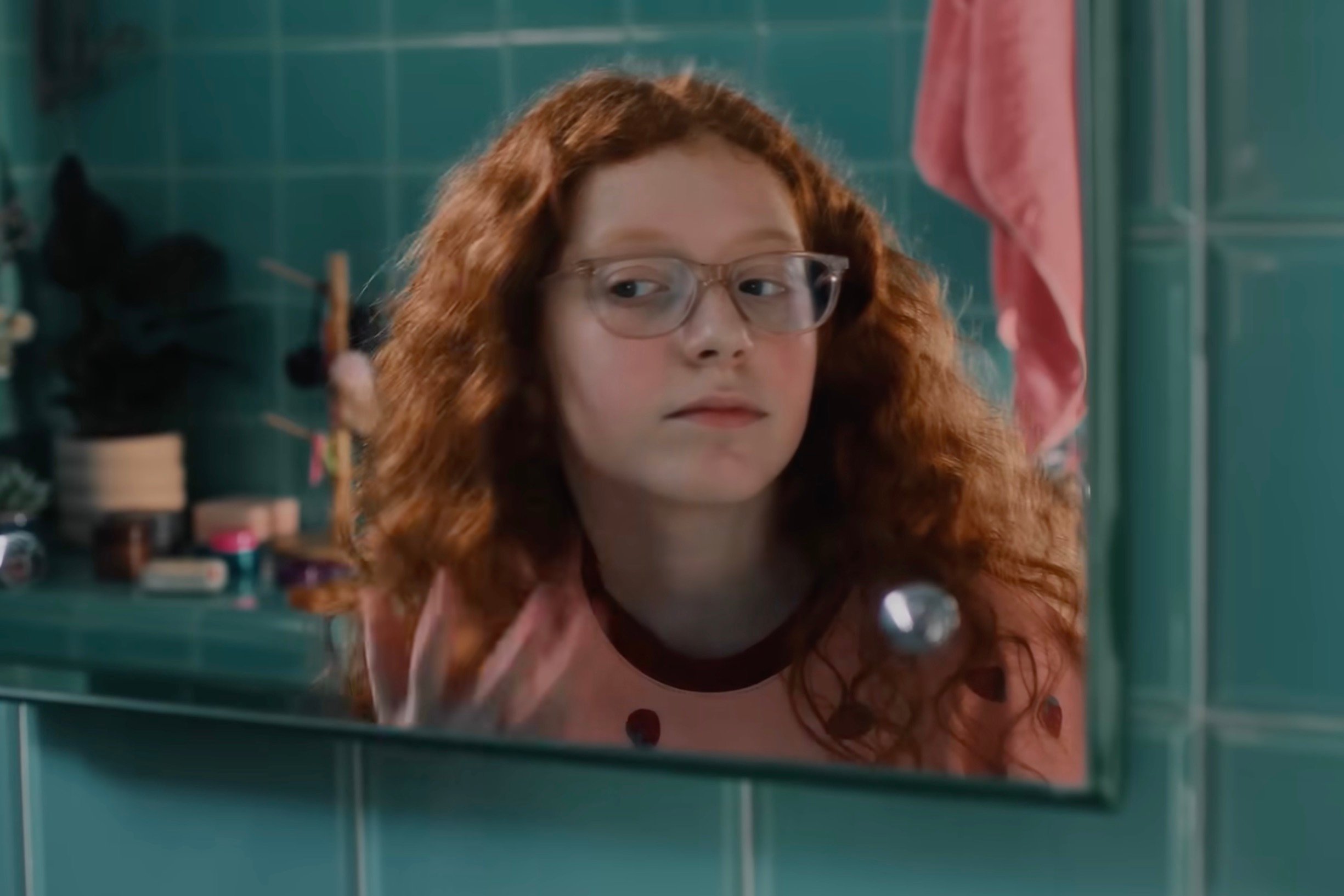 A young girl with curly red hair and glasses looks at herself in a bathroom mirror, touching her hair. The bathroom features teal tiles and toiletries on the counter behind her 