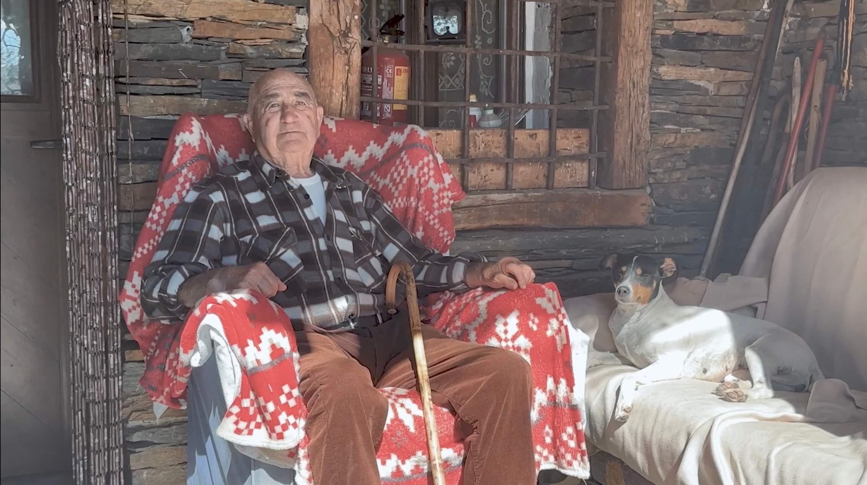 Elderly man sitting in an armchair with a walking stick and a red patterned blanket on the porch of a stone building, with a small dog resting beside him 