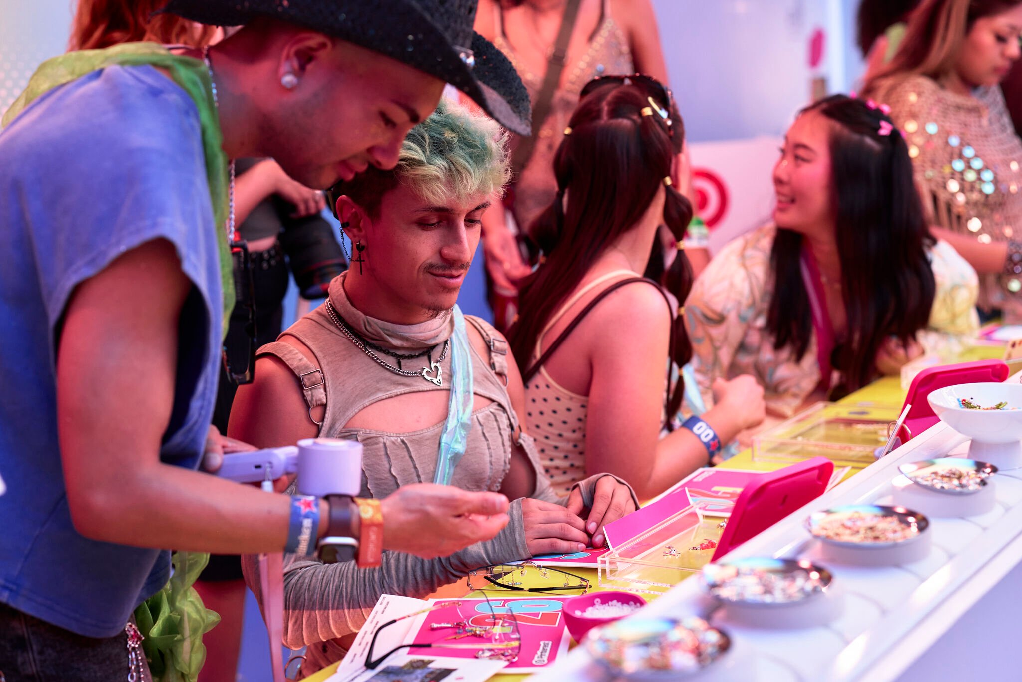 Two young people side by side at a crafting station, assembling custom charm accessories from bowls of beads and small decorative pieces laid out on pink trays along a yellow table 