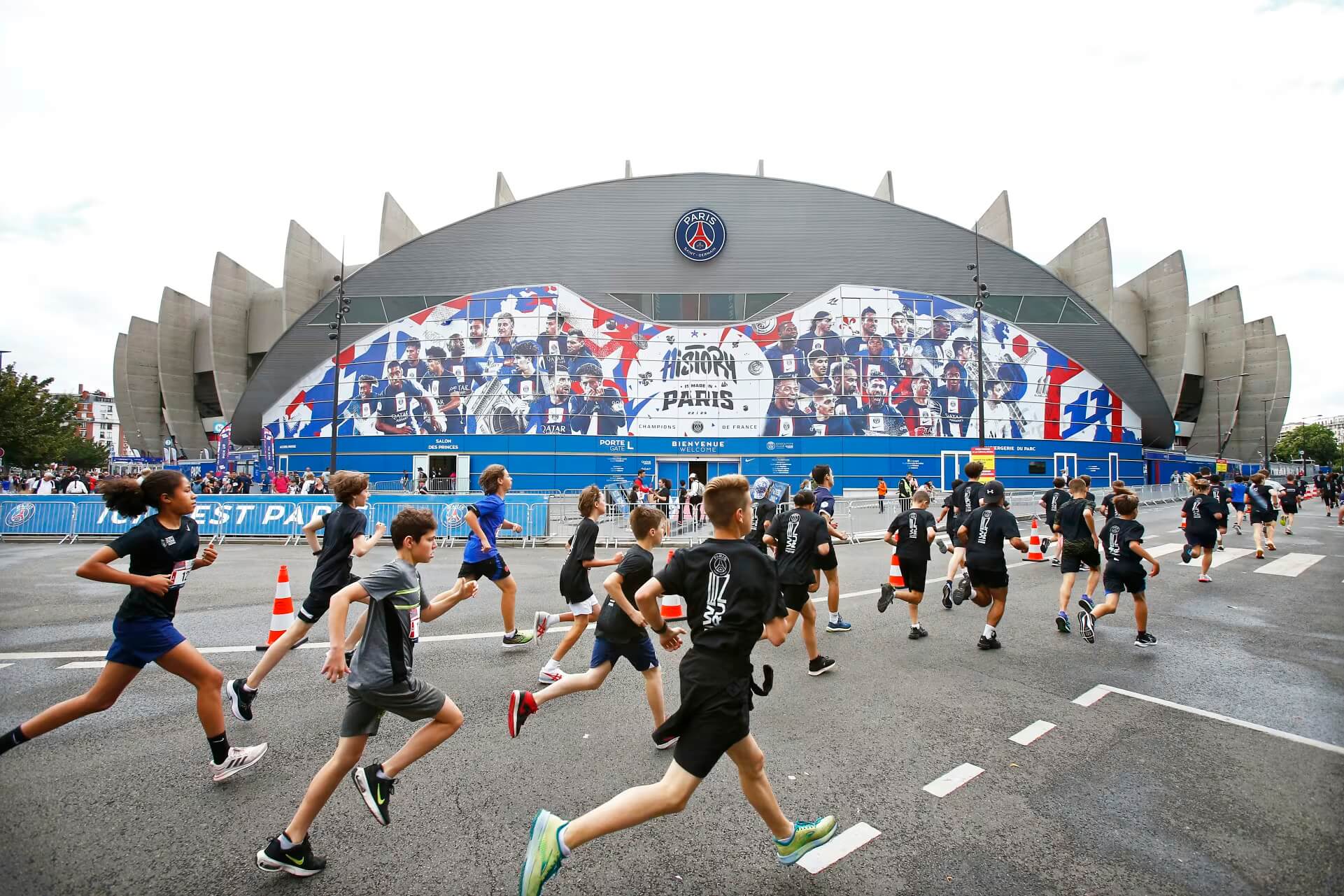 A large crowd of young runners wearing PSG and Nike gear races along a Parisian street, with the Parc des Princes stadium visible in the background 
