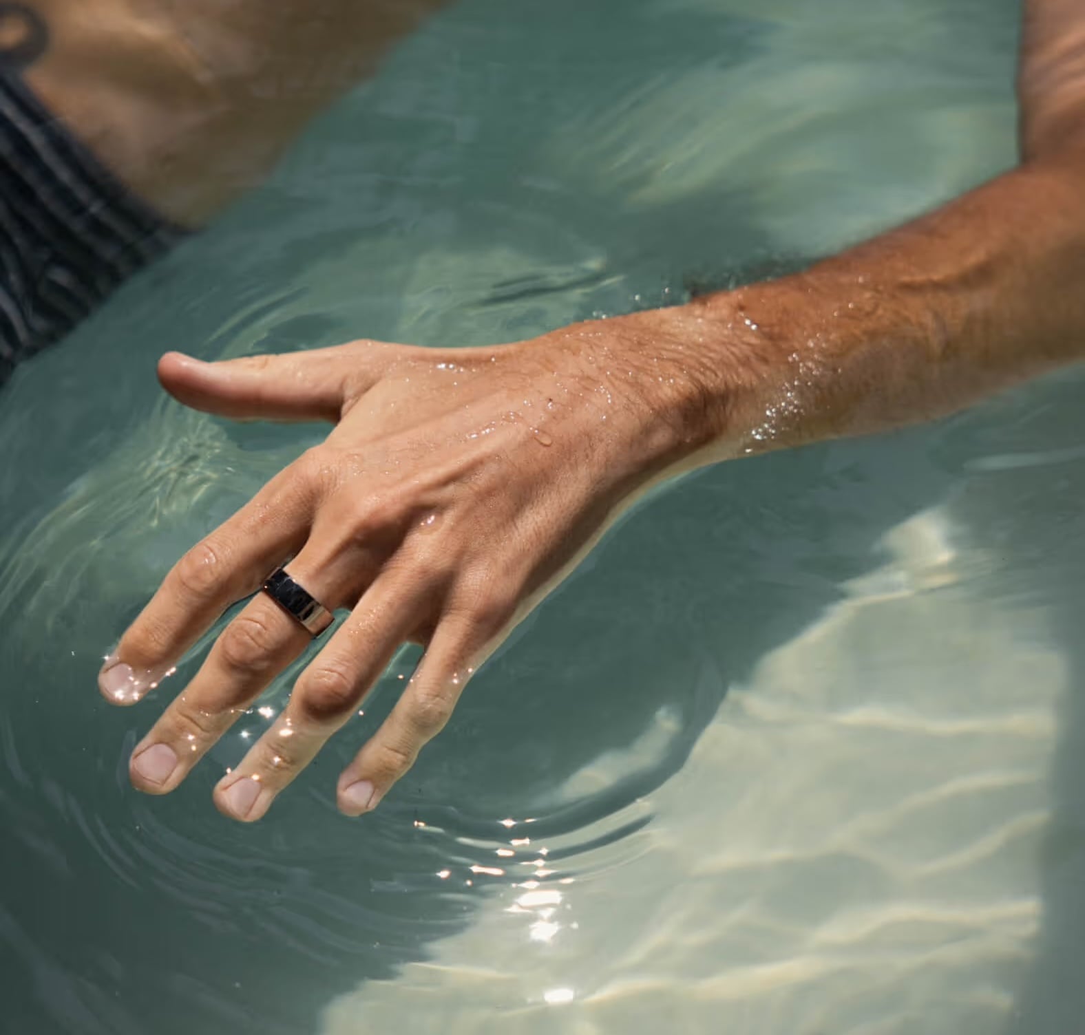 A man's hand wearing a dark smart ring, fingers resting just below the surface of clear blue-green pool water 