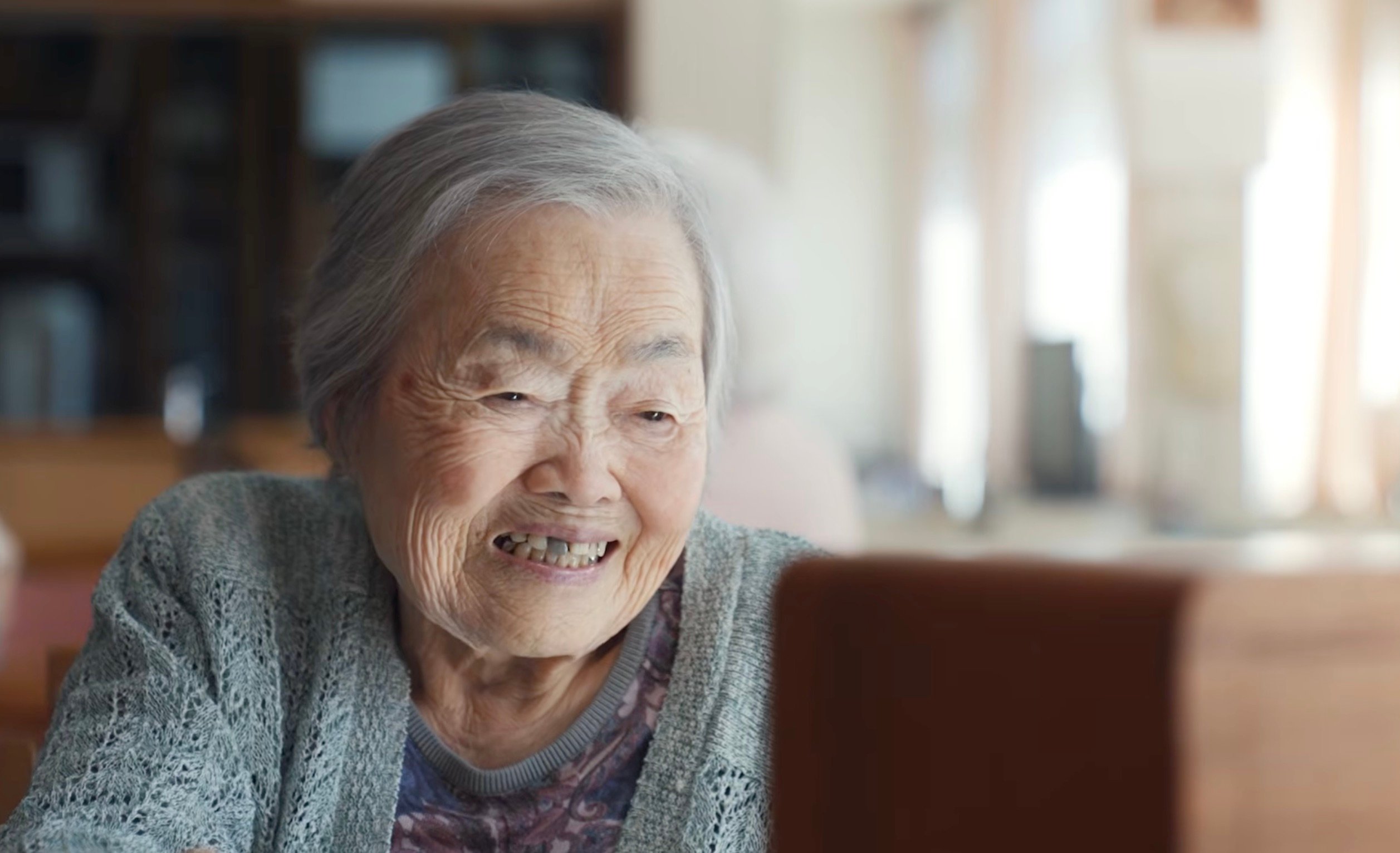 Elderly woman with short gray hair smiling warmly while seated in a softly lit care facility common area 
