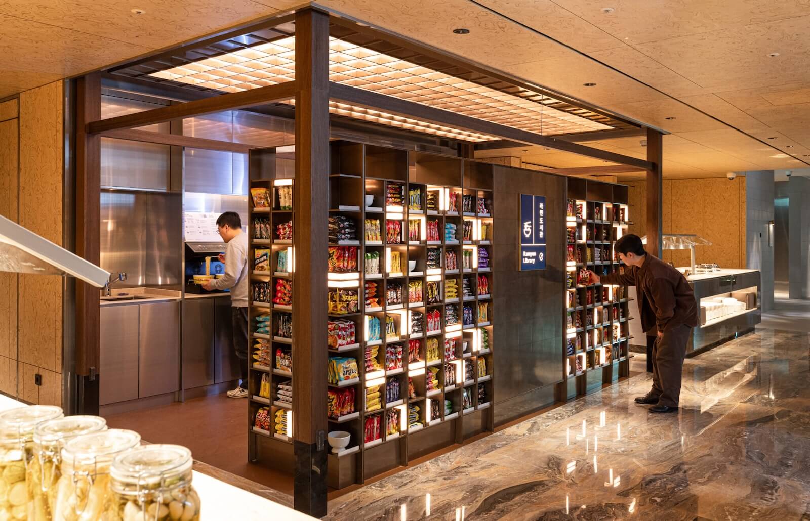 Backlit shelving units displaying dozens of colorful instant ramen packages in a modern airport lounge, with passengers browsing the selection at Korean Air's Ramyeon Library station 
