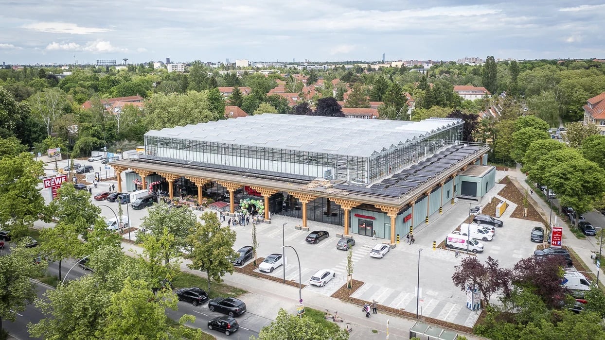 Aerial view of the REWE Green Farming supermarket in Berlin-Lankwitz, a long single-story timber building topped by a large glass rooftop greenhouse, with solar panels along the perimeter, a paved parking lot in front, and residential rooftops and tree canopy stretching to the city skyline behind 