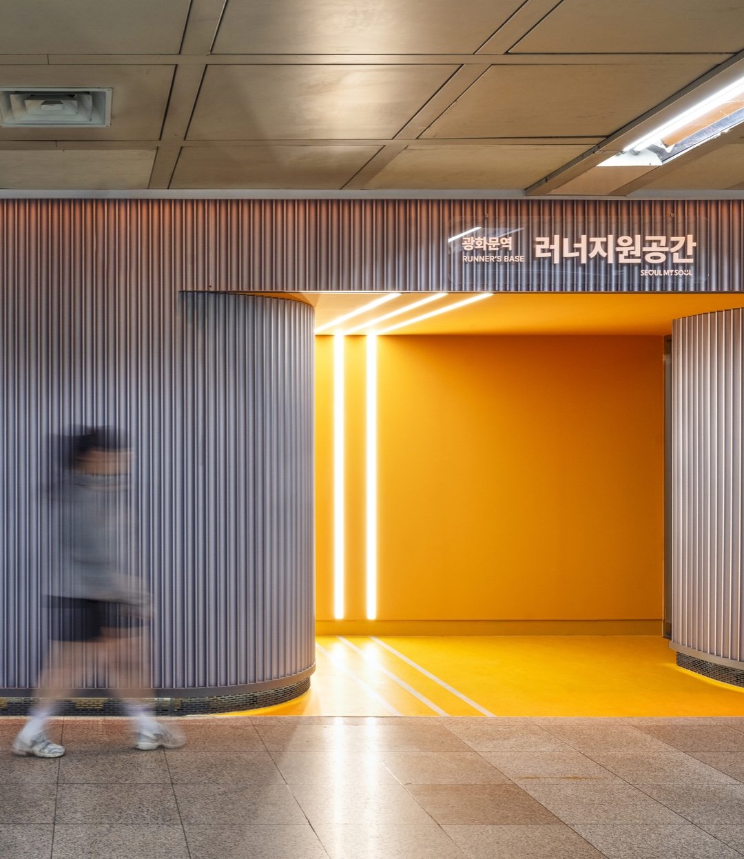 Motion-blurred person walking toward the entrance of the Runner’s Base at Gwanghwamun Station in Seoul, featuring a bright yellow interior with vertical light strips and curved gray walls 