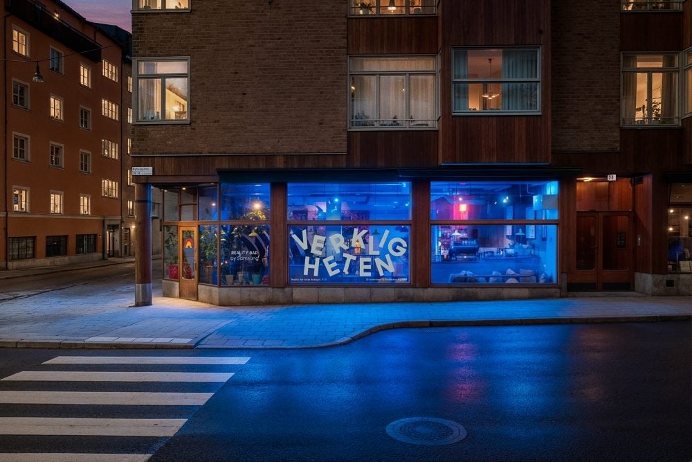 A street-corner view of Samsung’s Verkligheten pop-up reality TV bar in Stockholm, shown at dusk with blue interior lighting glowing through large front windows 