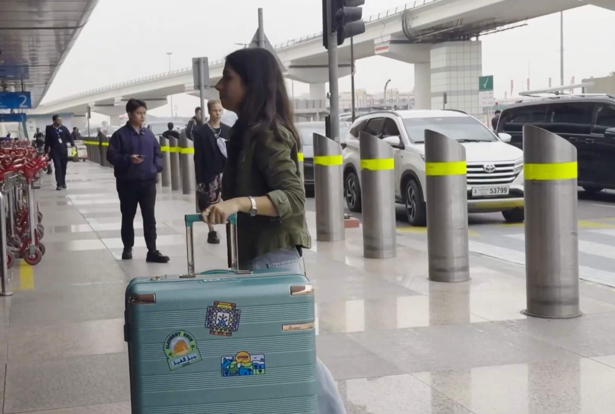 A young woman with long dark hair walks through an airport drop-off area, pulling a teal hard-shell suitcase decorated with travel stickers, including one that says 