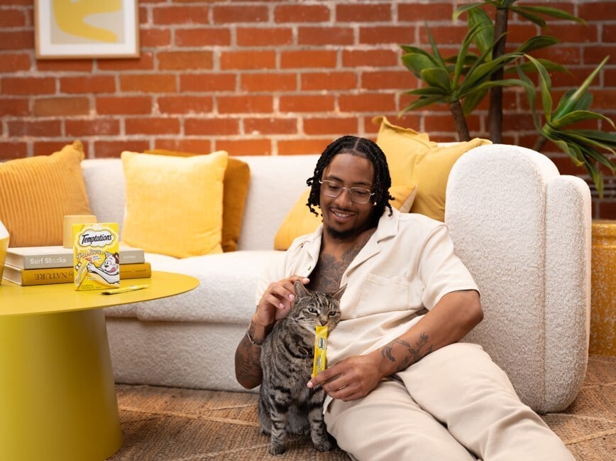 Man sitting on the floor against a cream sofa, smiling as he pets a gray tabby cat and feeds it Temptations treats 