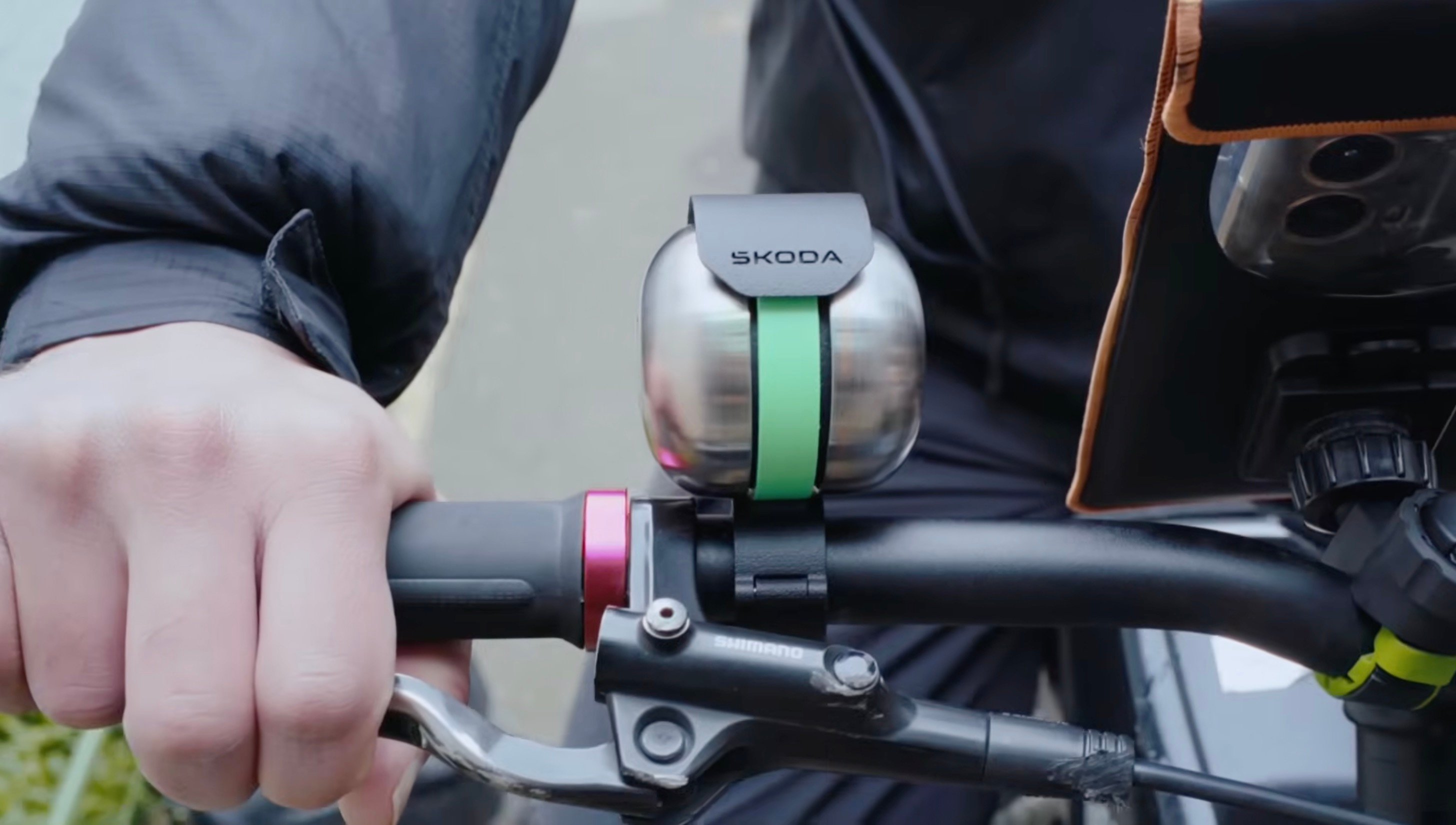 Close-up of the Škoda DuoBell bicycle bell mounted on a handlebar, showing its dual-dome metallic design with a green band and the Škoda logo on top 