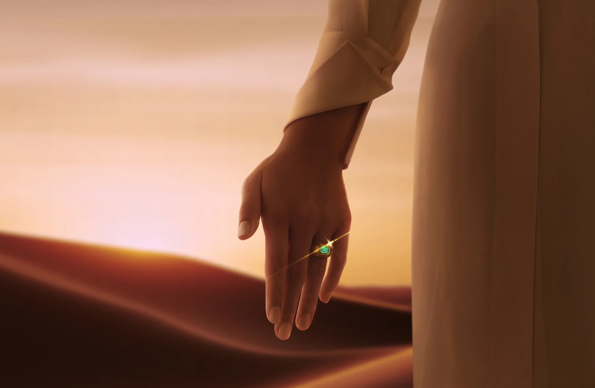 A person's hand extends against a warm, golden background, wearing a ring with a green gemstone 