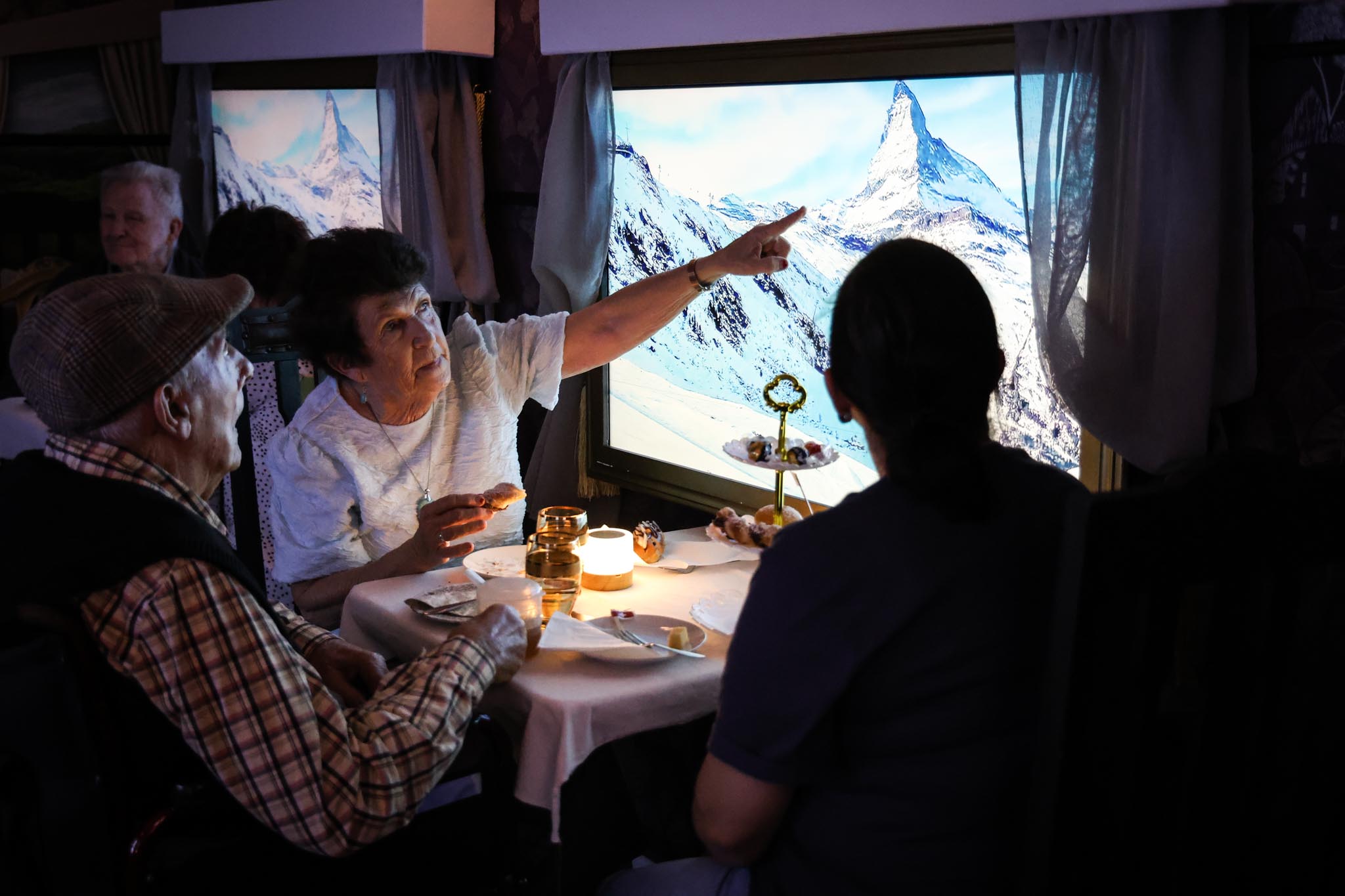 Three aged care residents seated at a candlelit dining table inside a mock train carriage, one woman pointing toward a large screen displaying the snow-capped Matterhorn in the Swiss Alps 