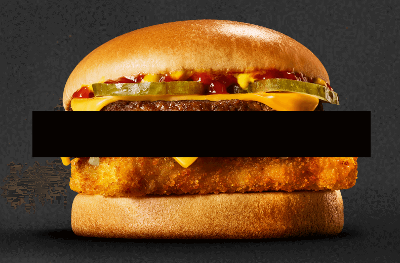 McDonald’s Surf ’N’ Turf burger with a beef patty and crispy fish fillet stacked with cheese, pickles, and sauce in a bun, partially obscured by a black 