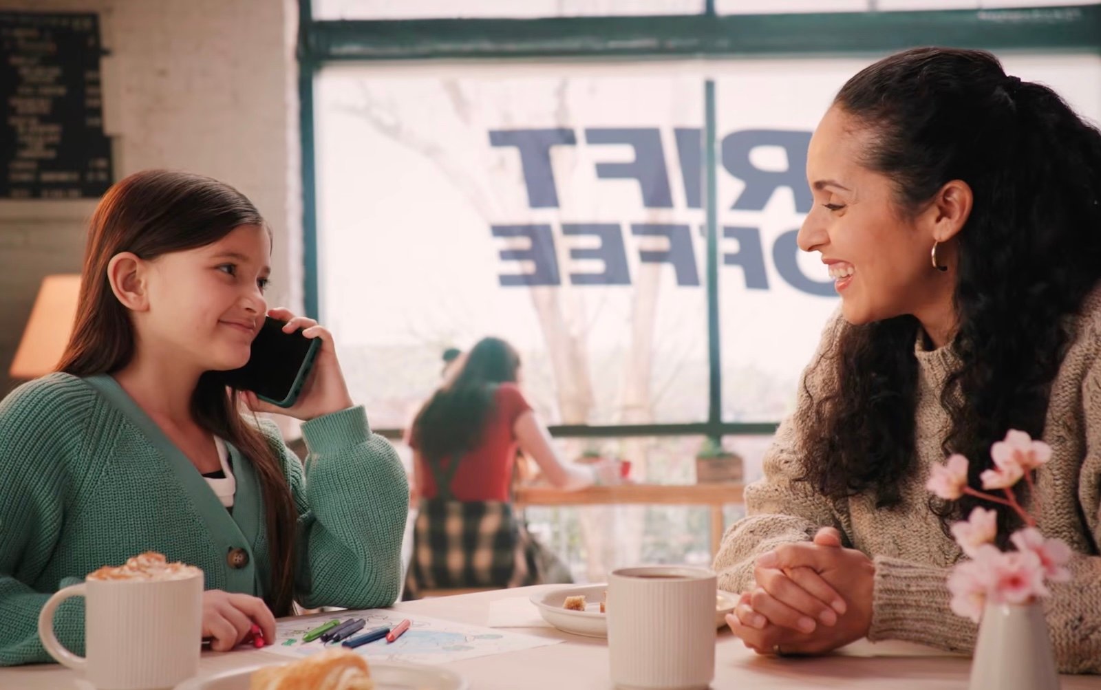 A young girl holding a smartphone to her ear smiles at a woman sitting across from her at a café table, with coffee cups, pastries and crayons on the table and a large window in the background 