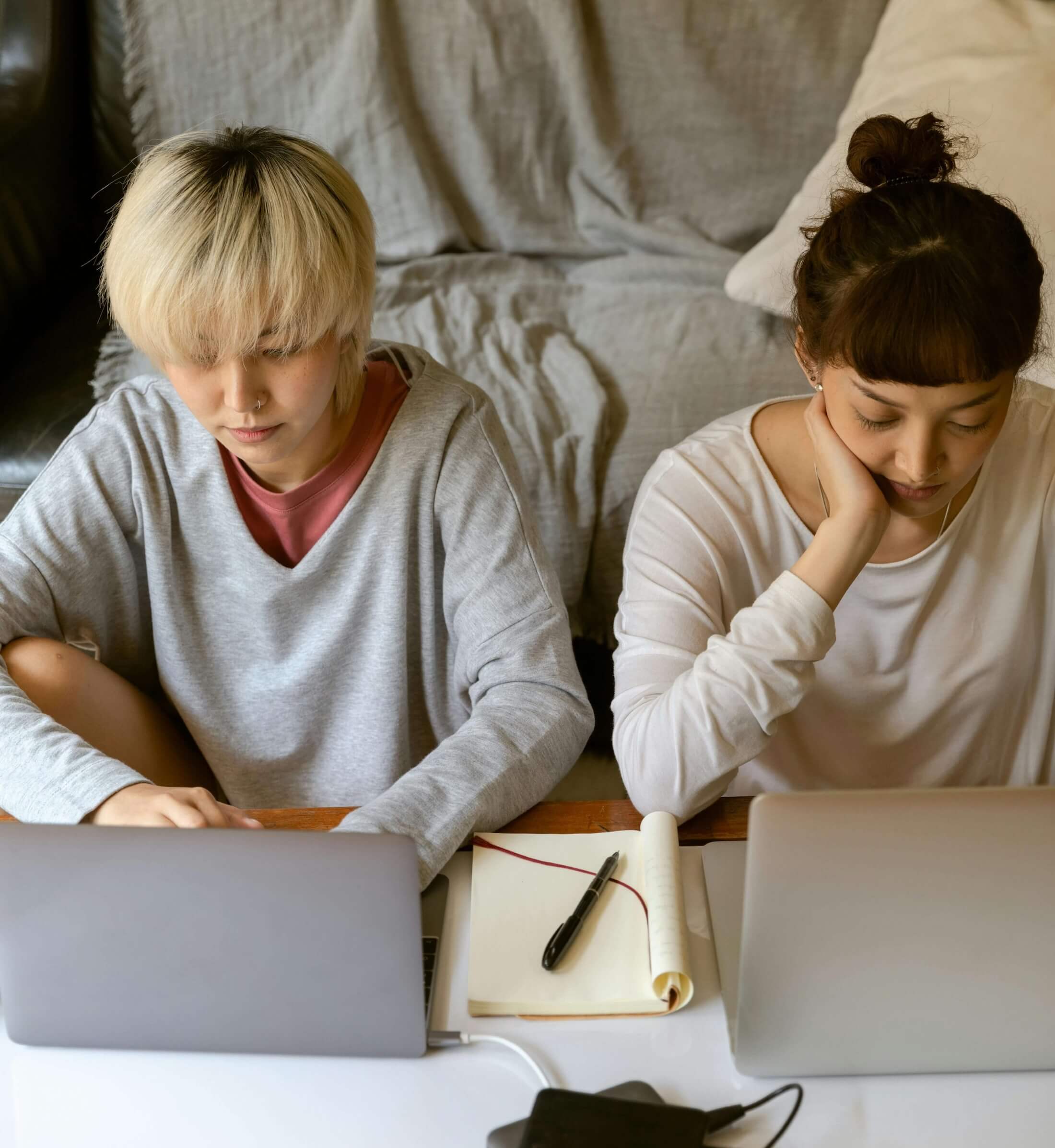 Two young people sitting side by side on the floor in front of a couch, each working on a laptop with a notepad and pen between them 