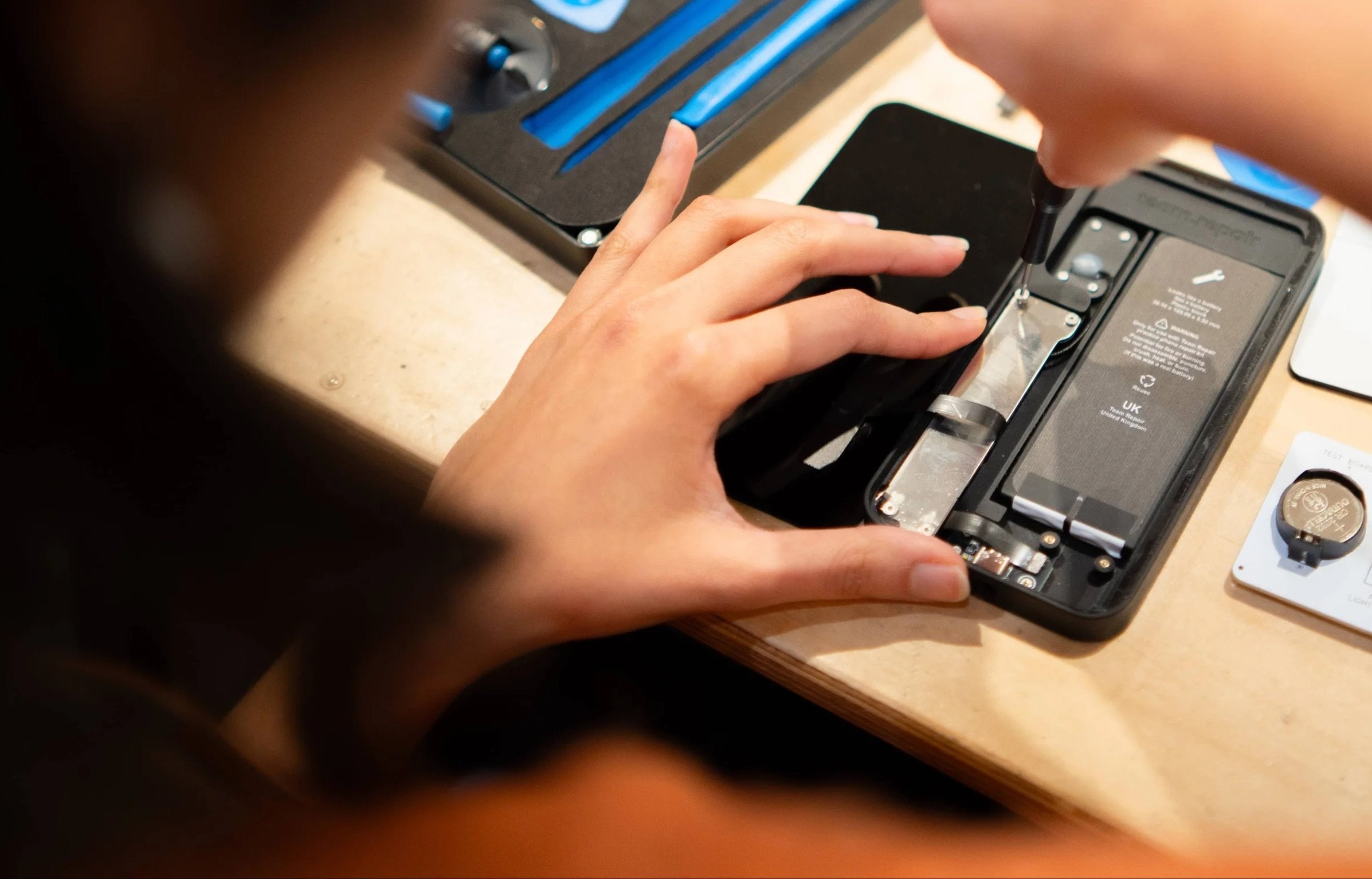 Close-up of two hands using a small screwdriver to work on the opened back of a black practice smartphone, with a battery and circuitry visible inside and a blue toolkit in the background 