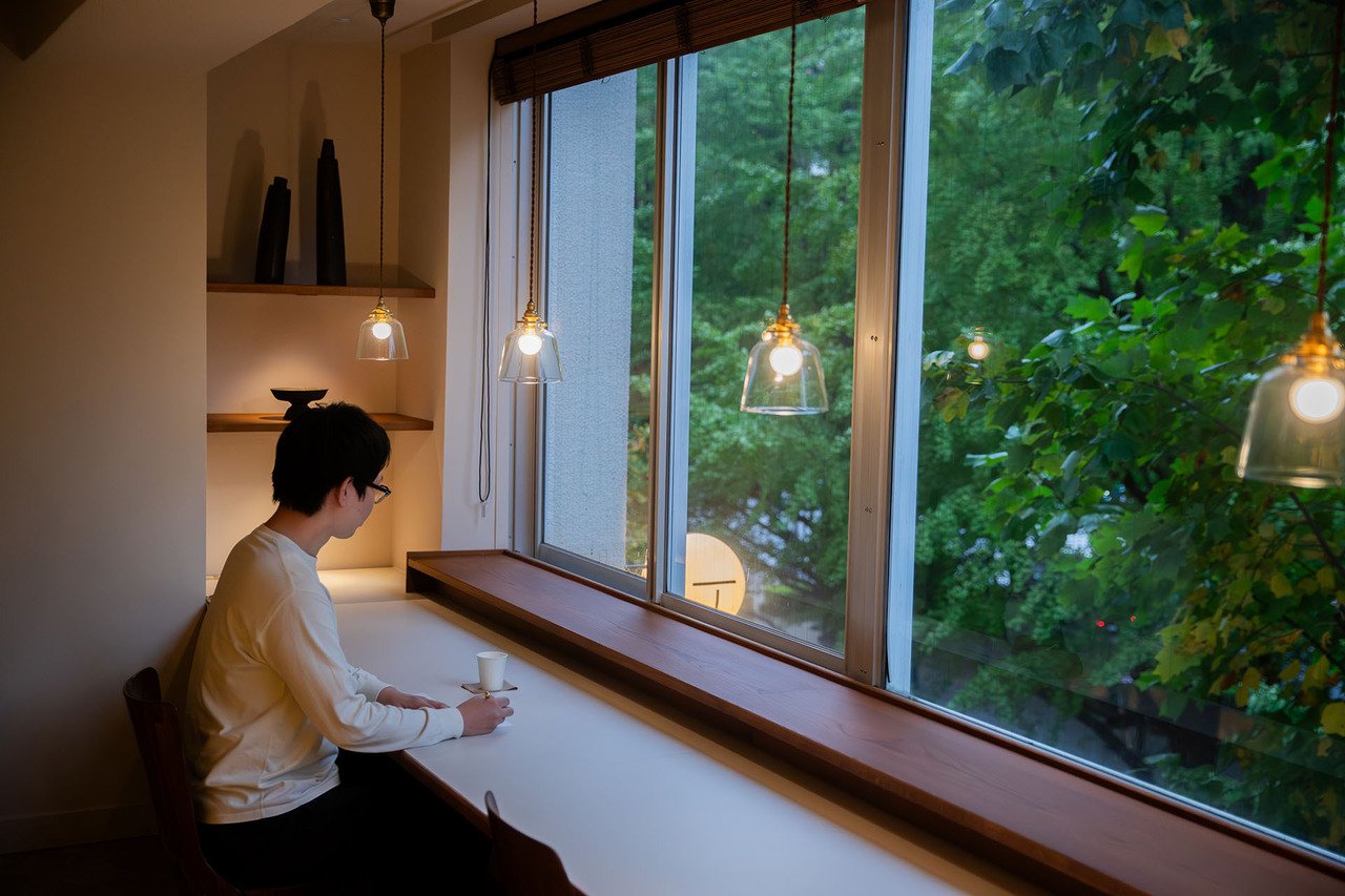 A person sits alone at a long counter by a large window, holding a cup in a softly lit room with pendant lights and a view of dense green trees outside 