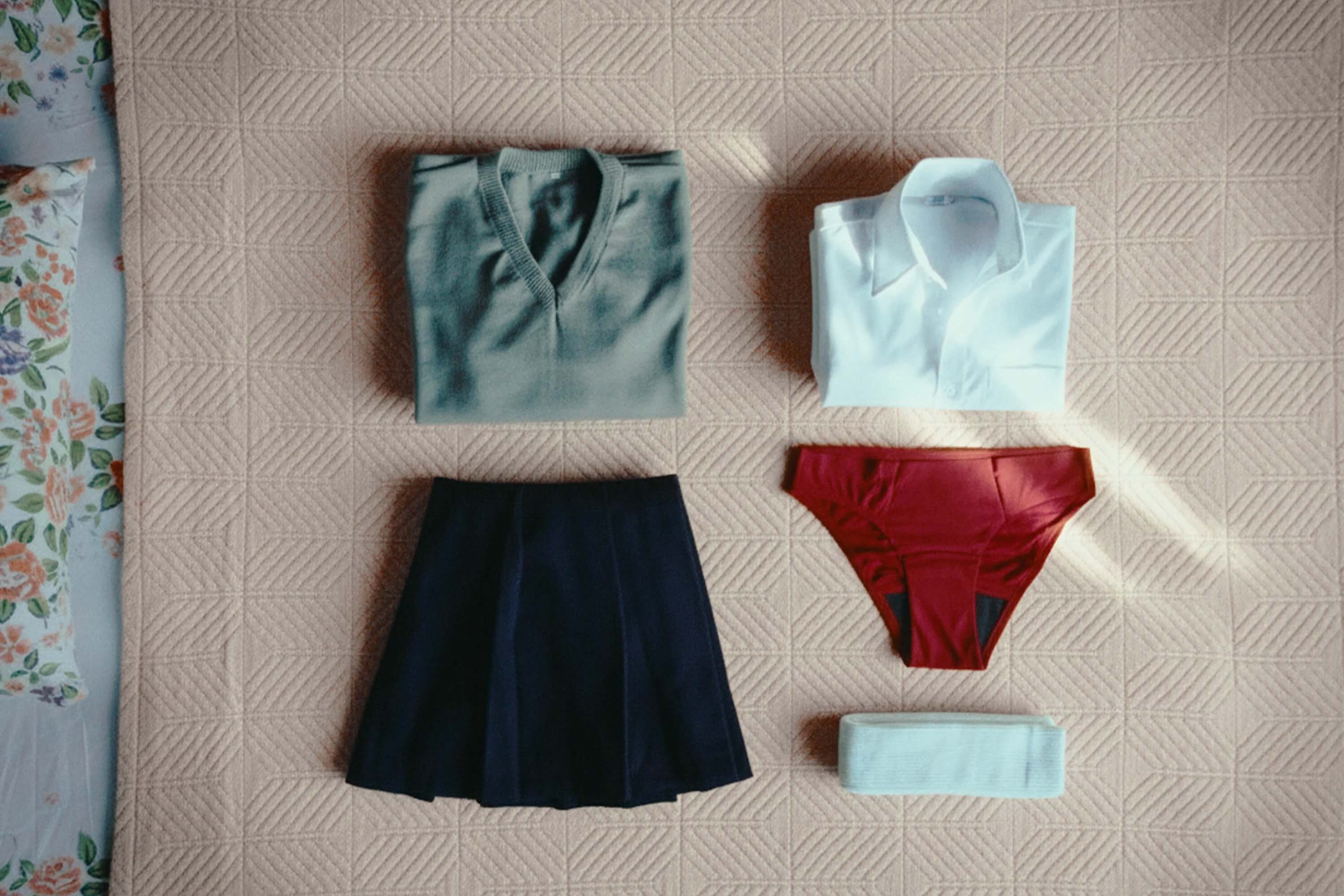 School uniform components laid flat including navy pleated skirt, gray cardigan, white polo shirt, burgundy period underwear, and white socks on textured beige surface 