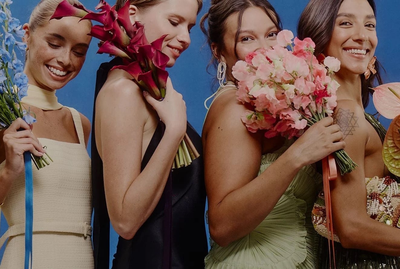 Four women in formal dresses stand close together against a blue backdrop, each holding a bouquet of flowers, smiling and looking down 