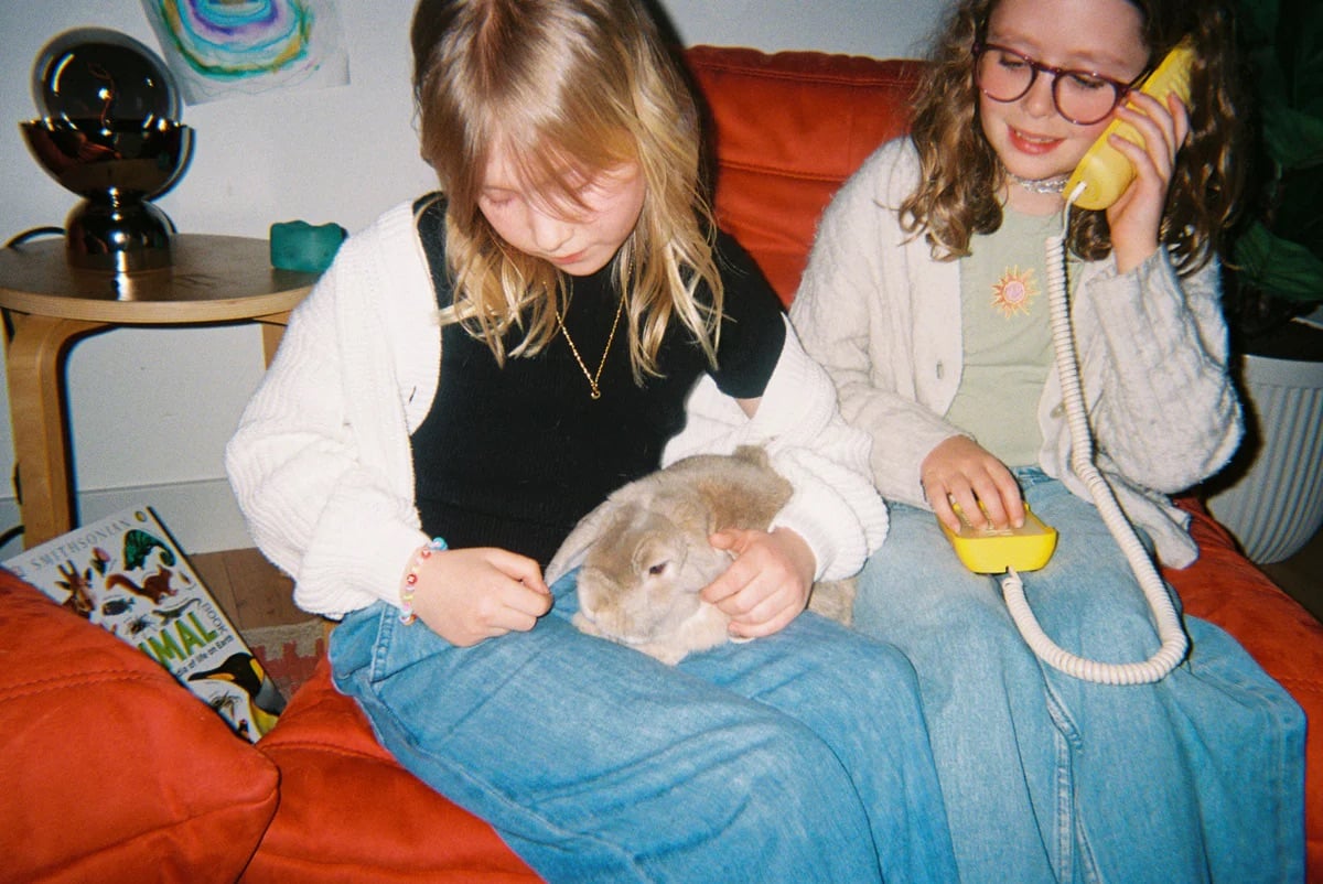 Two girls on a red couch; one pets a rabbit on her lap, while the other holds a yellow corded phone to her ear 