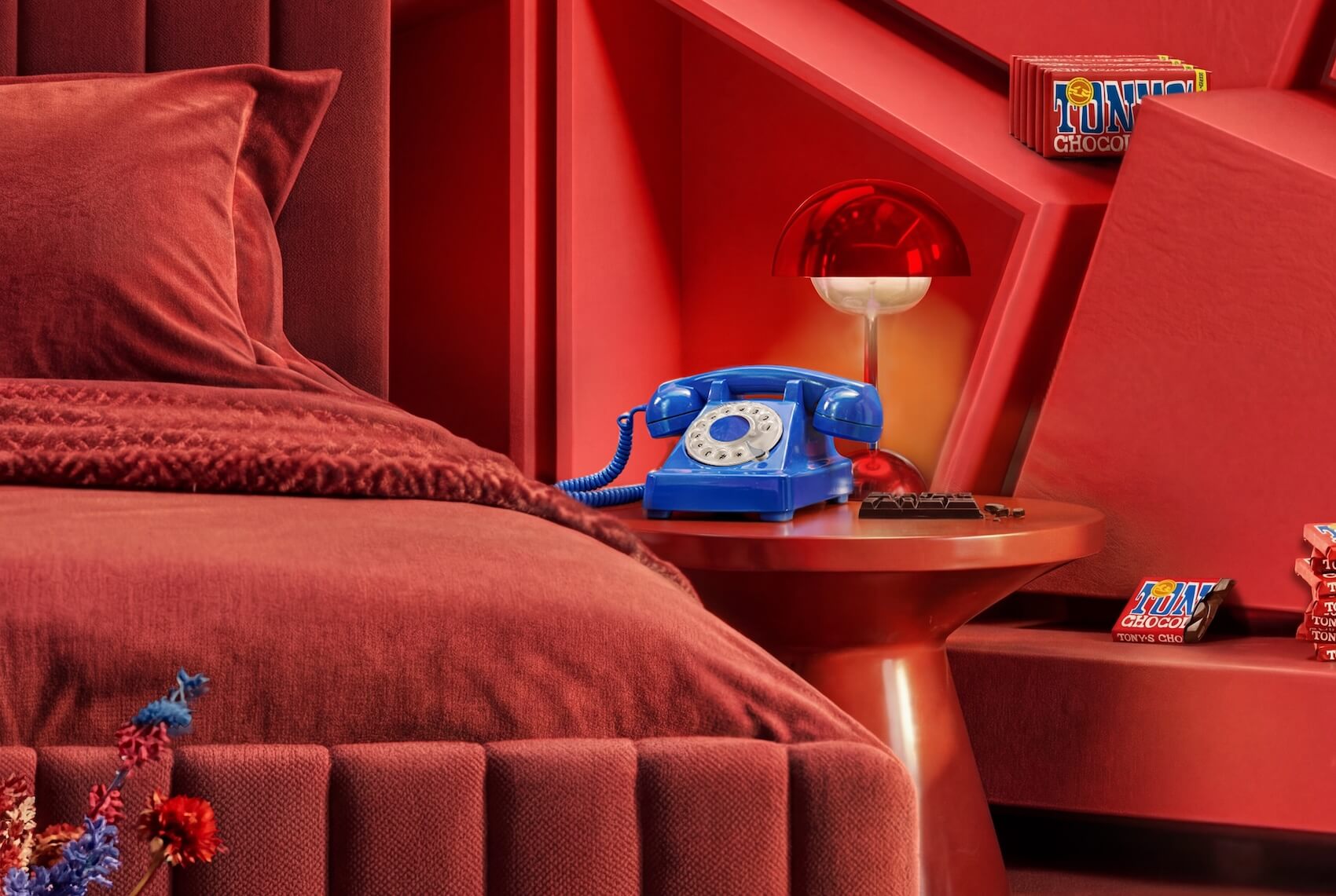 Red-themed hotel room with plush bedding, a bright blue rotary telephone on the nightstand, and Tony's Chocolonely chocolate bars displayed throughout 