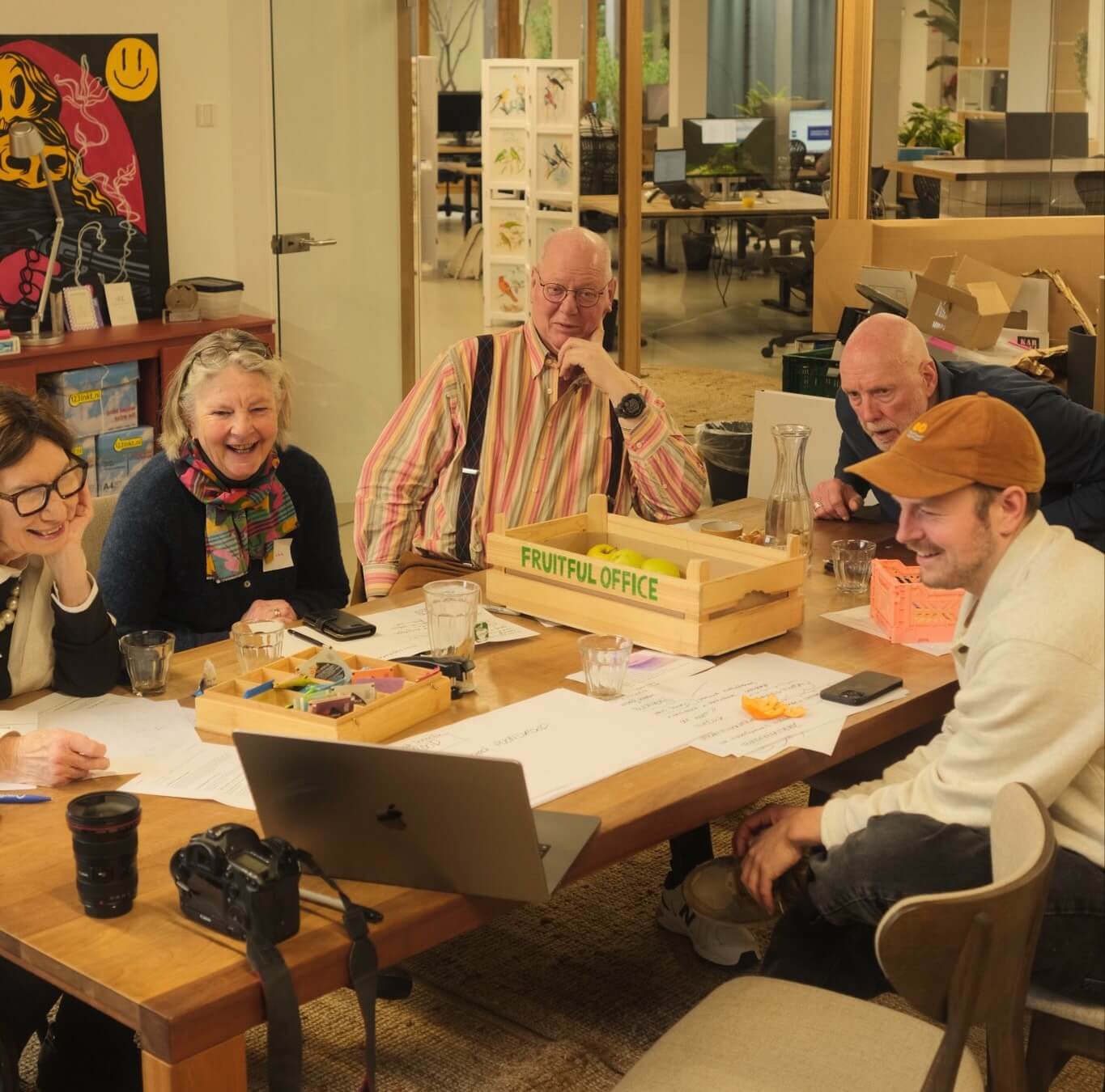 A group of older and younger adults laughing and collaborating around a table covered in markers, paper and laptops at a creative agency office 