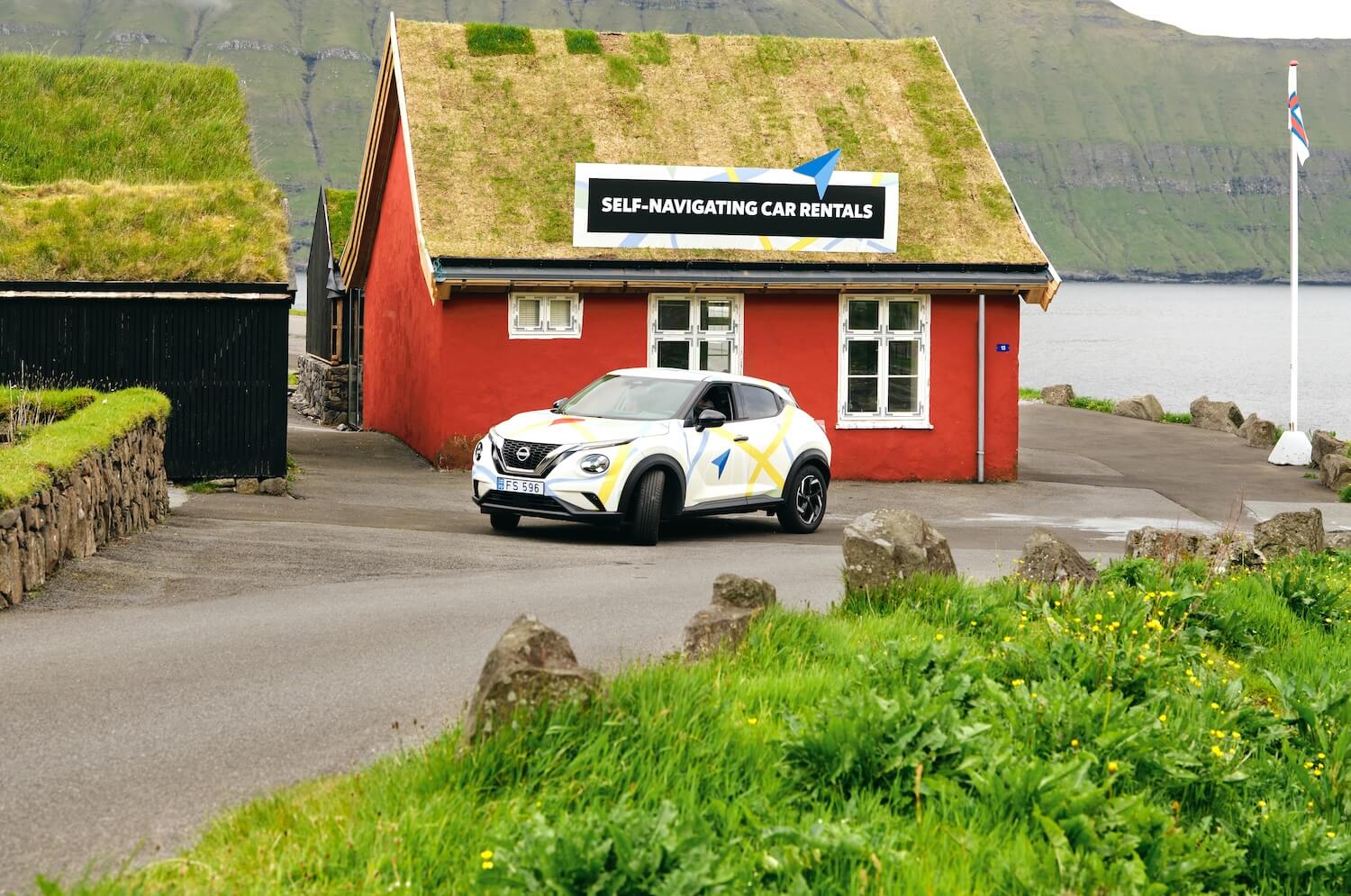 A rental car in front of a red building with a turf roof, labeled 'SELF-NAVIGATING CAR RENTALS' 