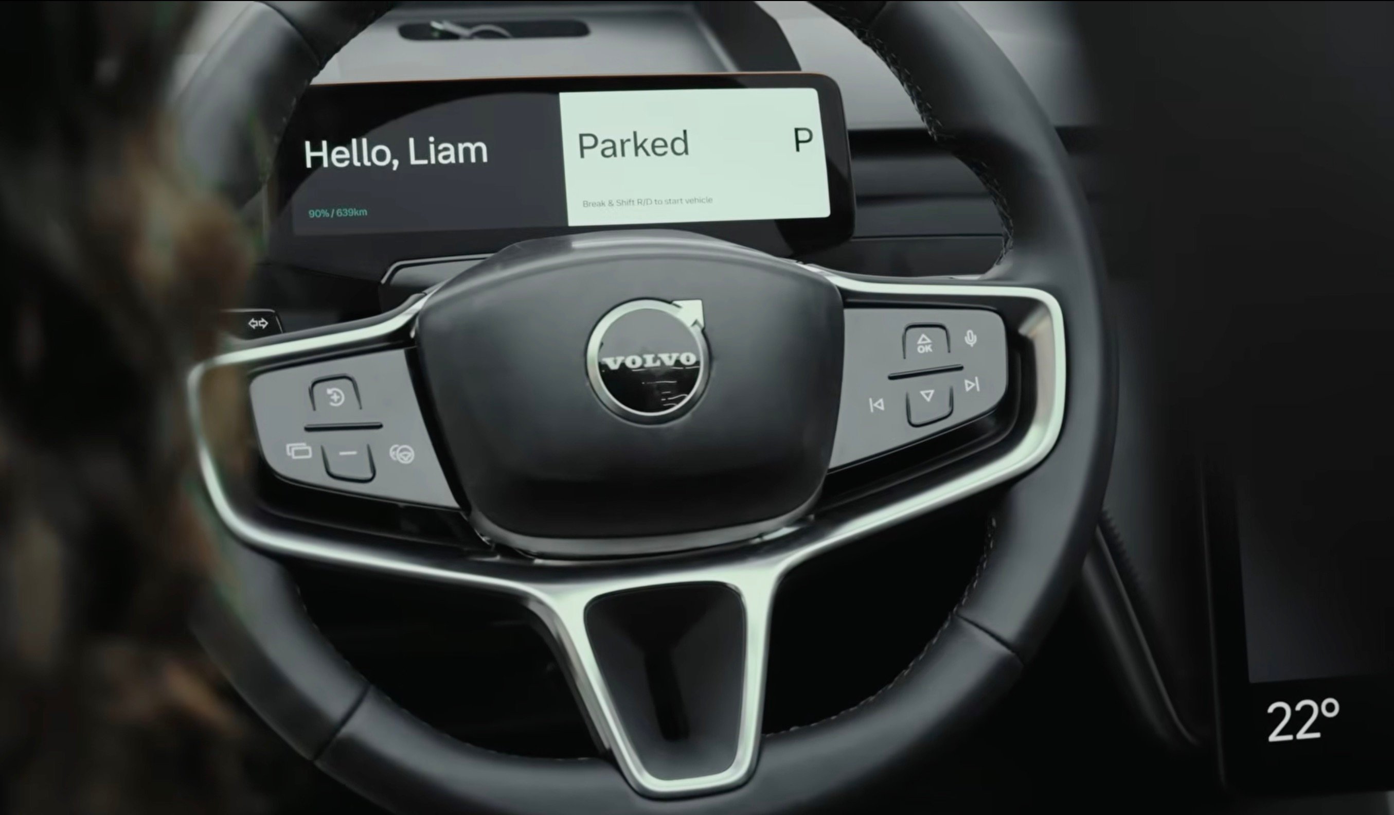Close-up view of a Volvo steering wheel and digital dashboard, showing a wide, minimalist display reading “Hello, Liam” and “Parked” 