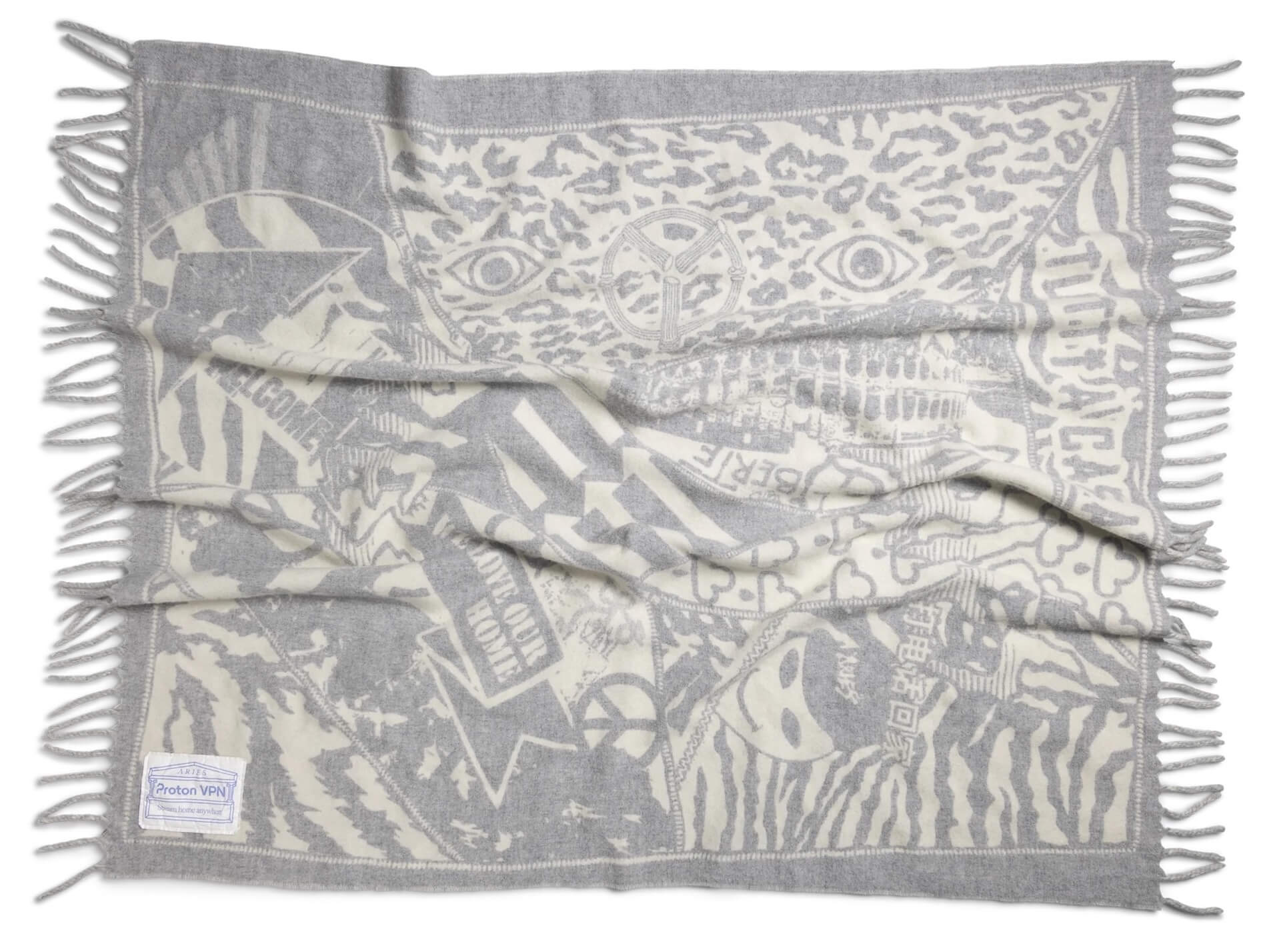 A gray-and-cream woven blanket with dense, collage-style graphics, including eyes, abstract patterns, text fragments and global iconography. Fringed edges run along all sides; a small fabric tag displays the Proton VPN and Aries branding 