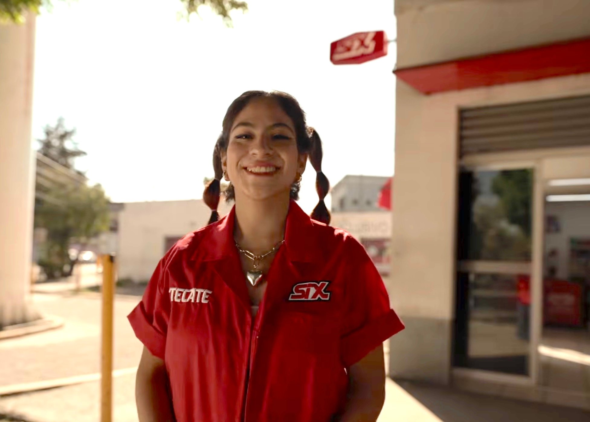 A young woman in a red Tecate-branded work shirt with a SIX logo patch smiles at the camera outside a Tiendas SIX convenience store on a sunny day 
