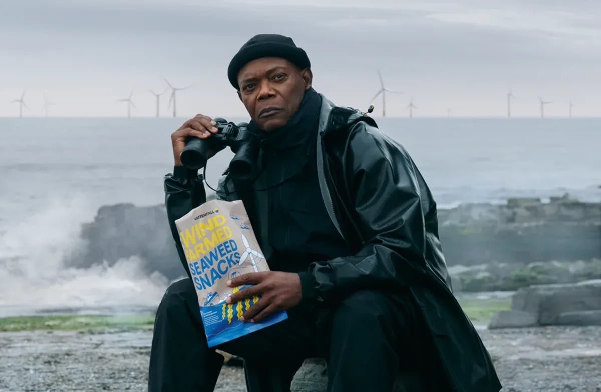Dressed in black outdoor gear, Samuel L. Jackson sits on a rock by the sea, holding a bag of “Wind Farmed Seaweed Snacks” and a pair of binoculars. Wind turbines are visible in the ocean background 