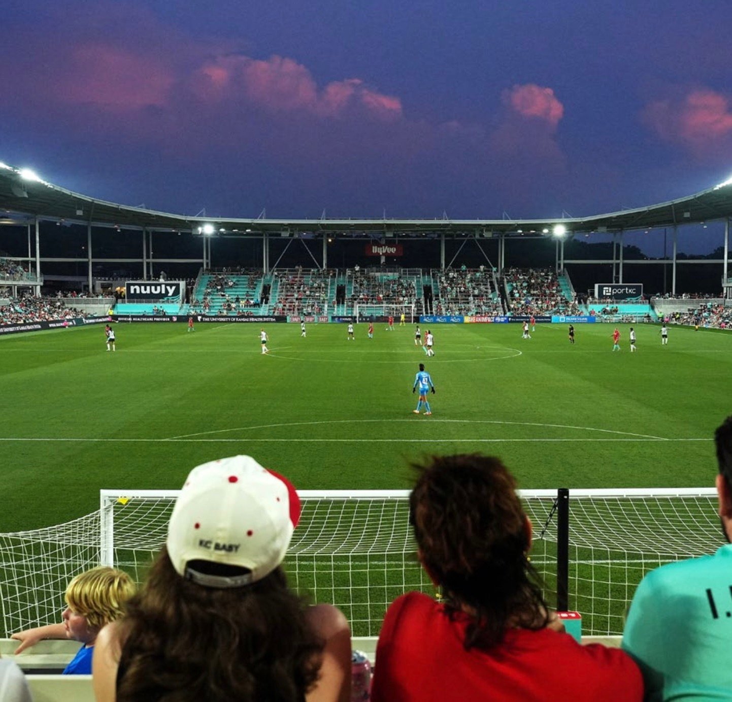 Evening match at CPKC Stadium with fans seated behind a goal, watching two women’s teams play on a brightly lit field. The stands are nearly full, and a purple-tinted sky hangs over the world’s first purpose-built venue for a professional women’s sports team 
